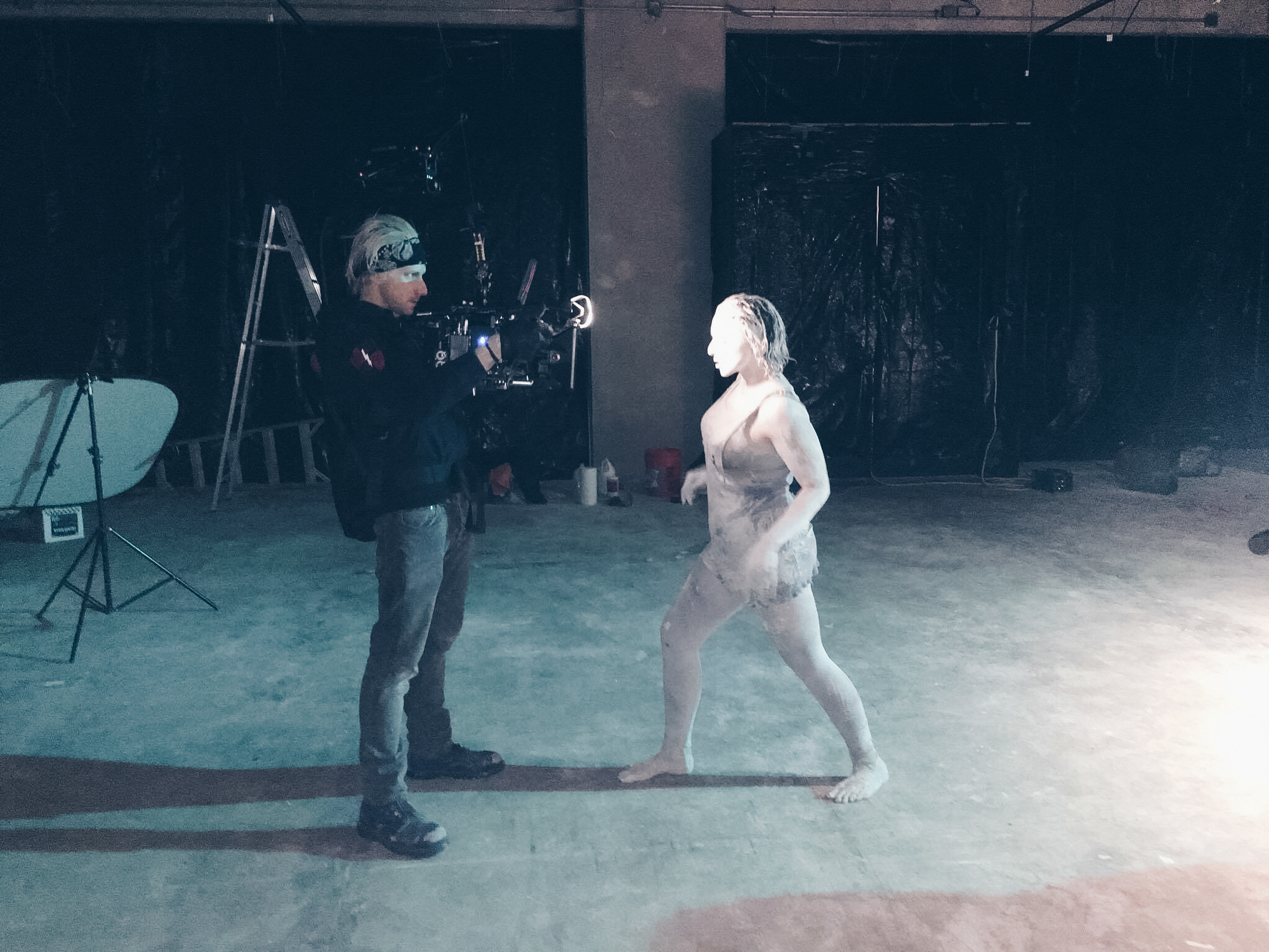 Man uses easyrig attached to camera to record a woman covered in white mud in dark room for a music video.