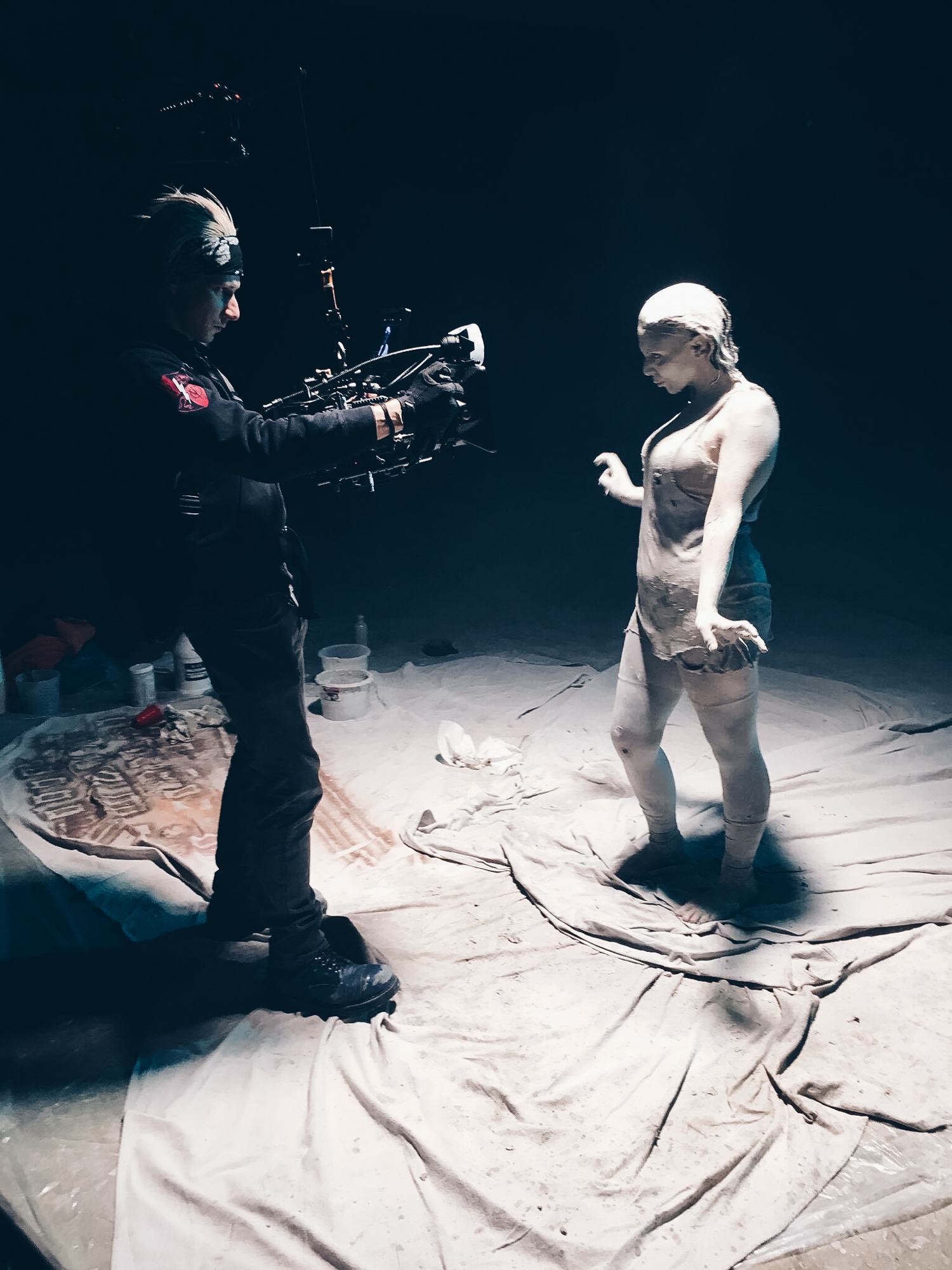 Man uses easyrig attached to camera to record a woman covered in white mud in dark room for a music video.