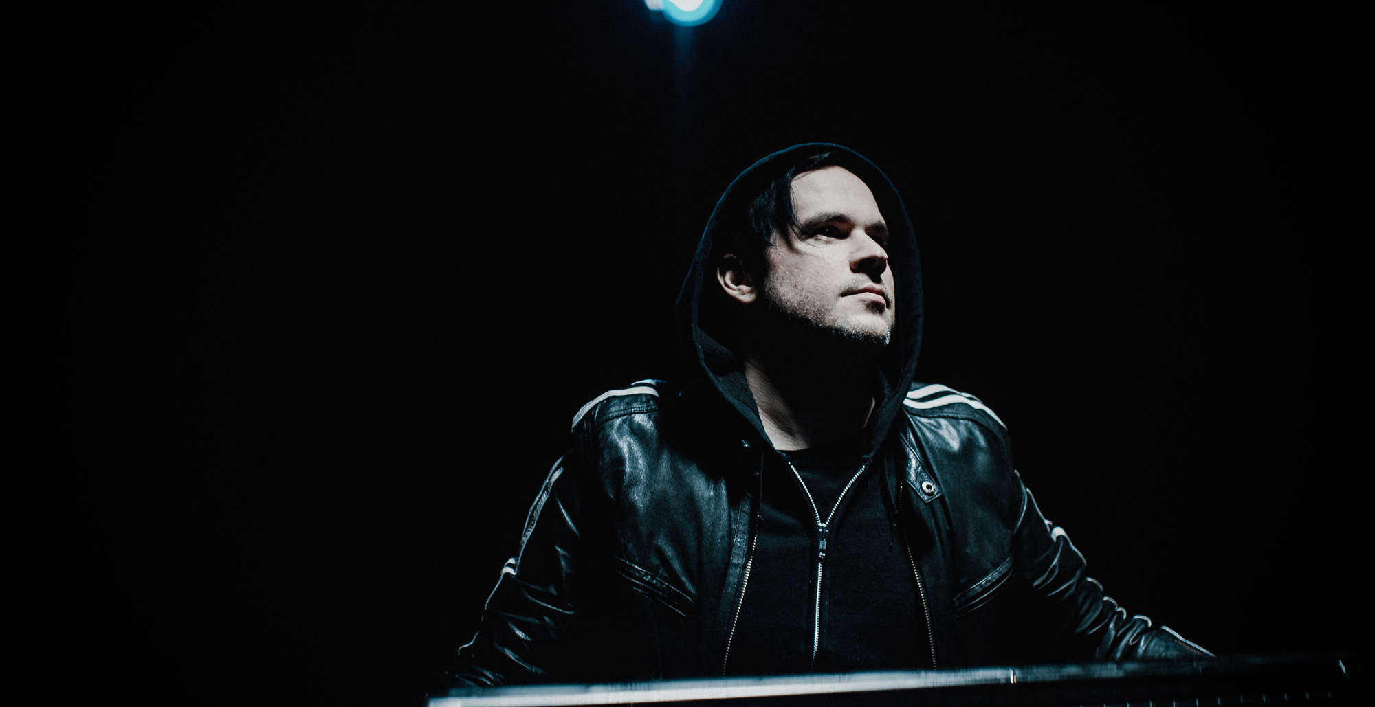 Man holds a keyboard with black leather jacket and hood in a black room lit by overhead light.