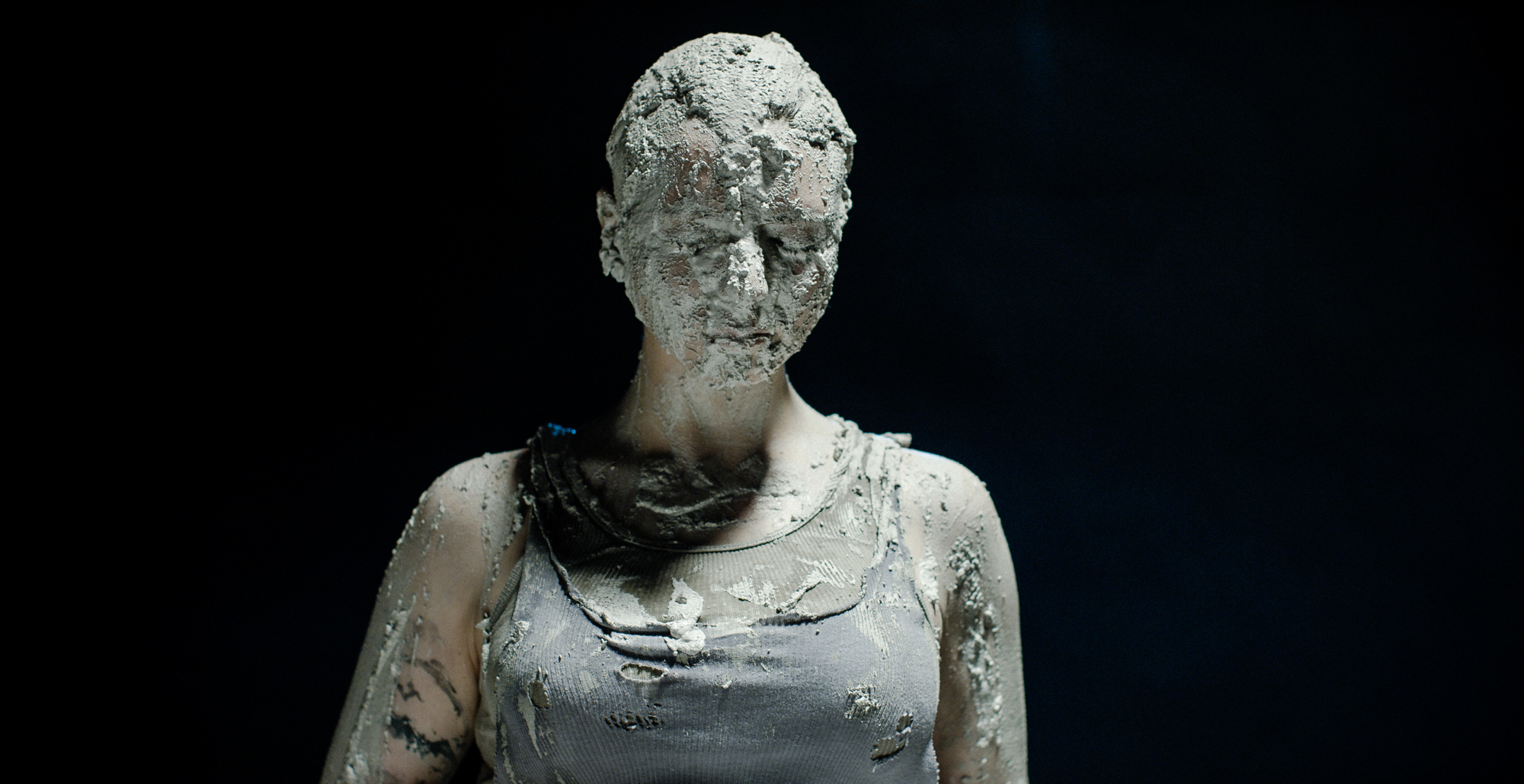 Woman covered in mud stand with eyes closed in a tank top.