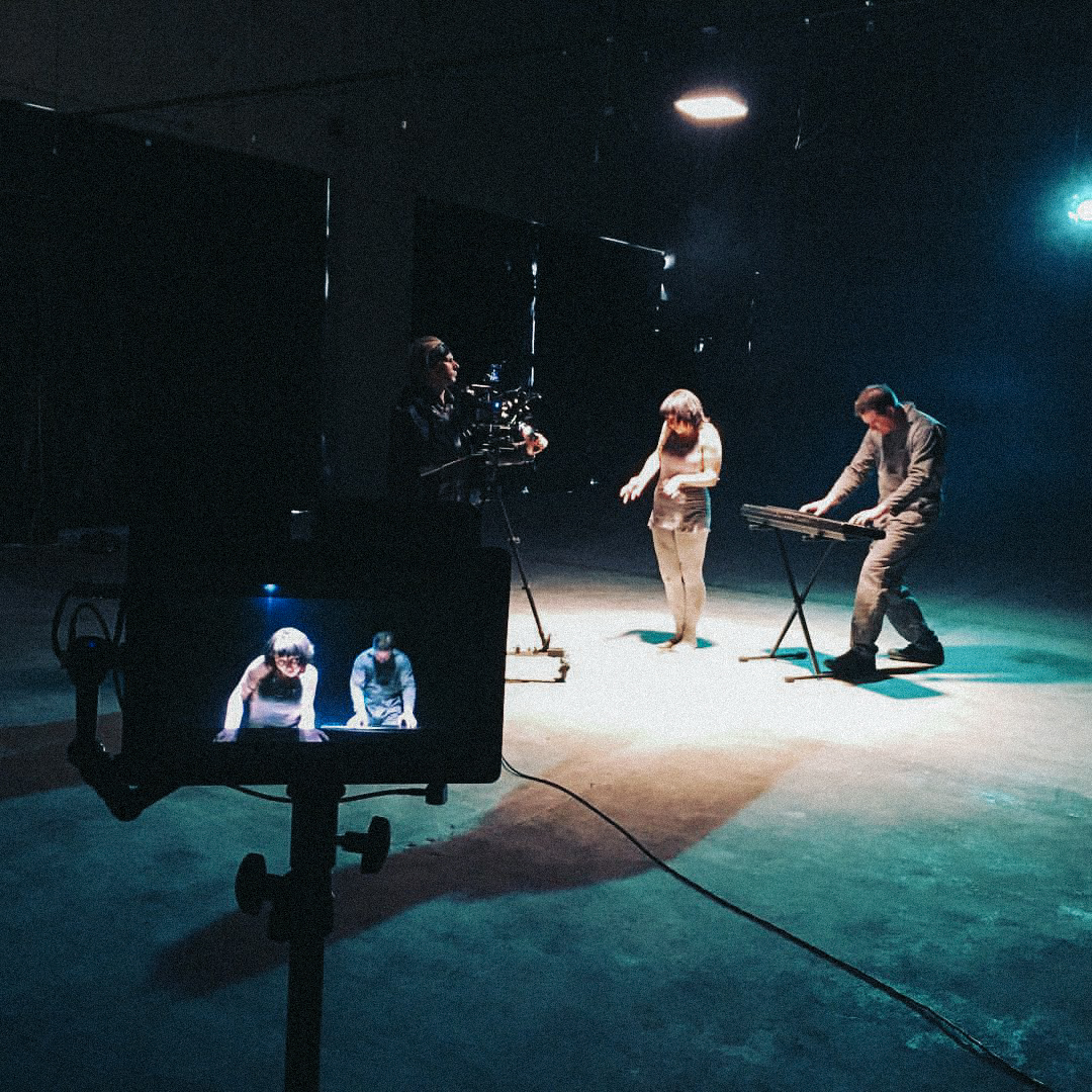 Man and woman perform for a music video as a cinematographer records under a light surrounded by darkness with a camera monitor in frame.