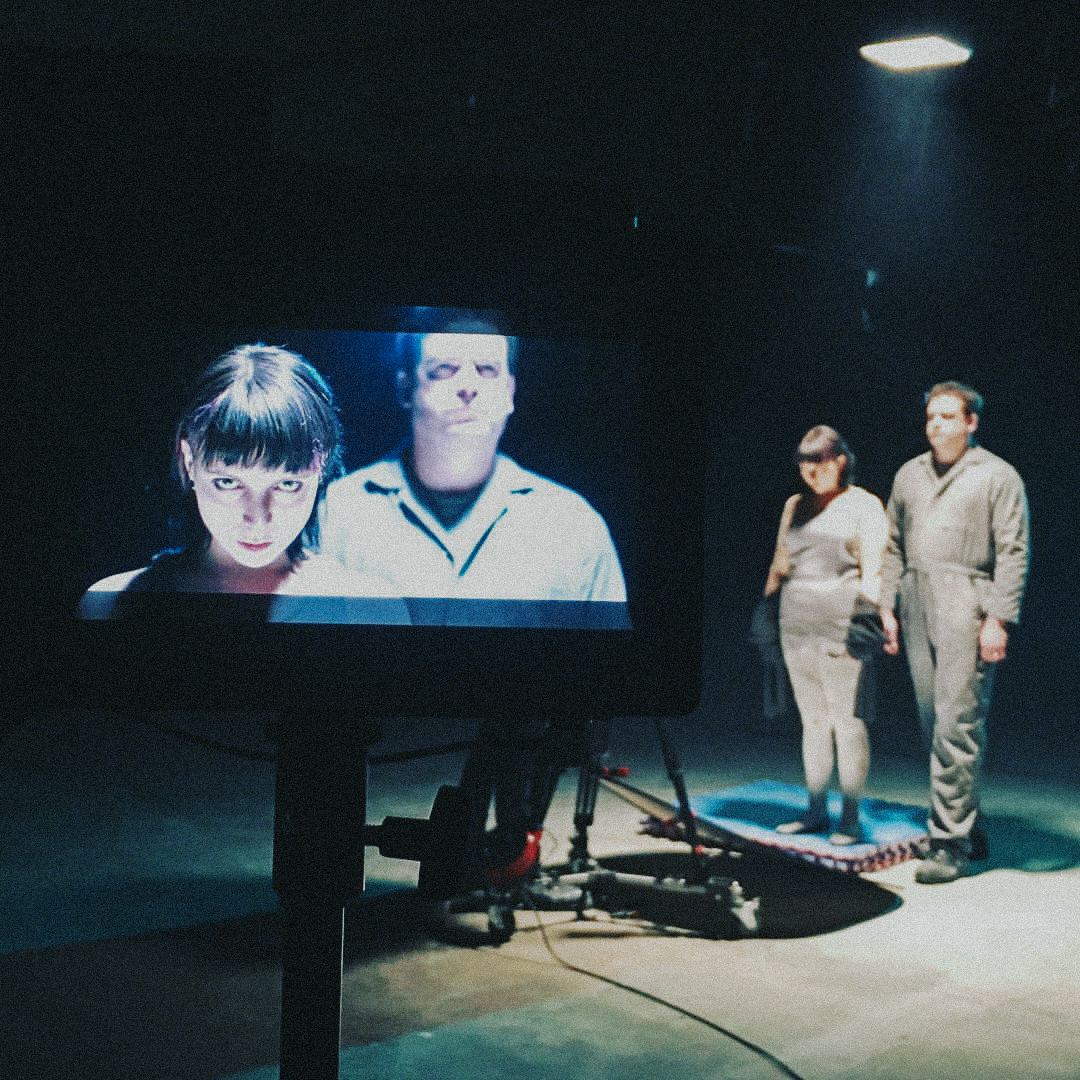 Man and woman dressed in overalls stand under a light surrounded by darkness with a camera monitor in frame.