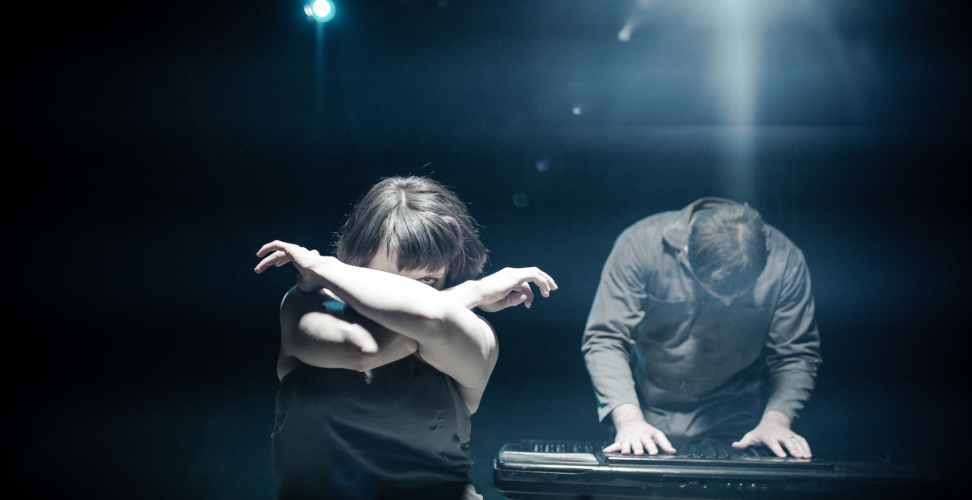 Woman crosses arms in face man plays keyboards in dark room.
