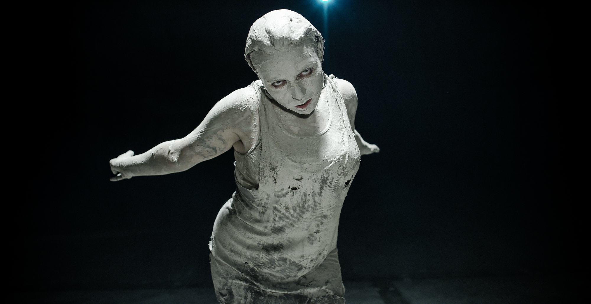 Woman covered in white clay stretches out to camera arms back.