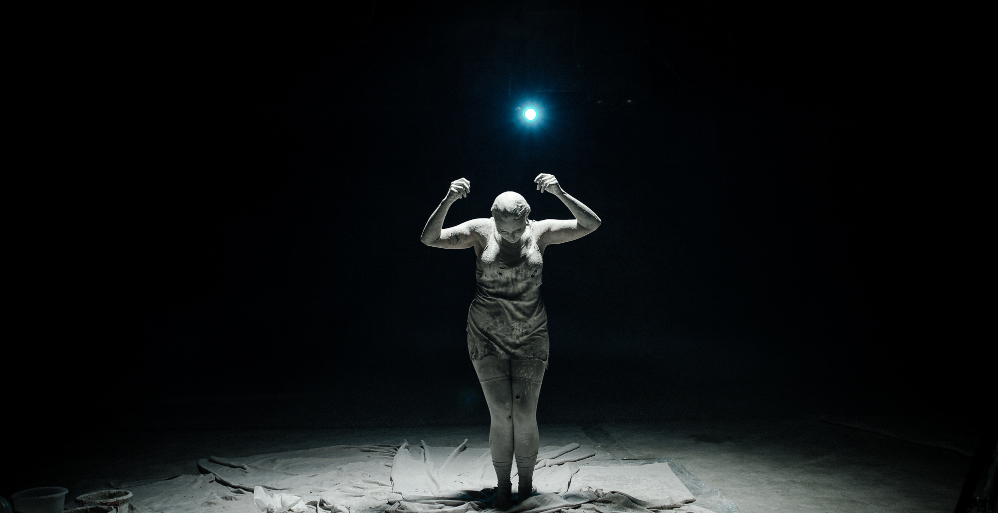 Woman covered in clay stands stretching arms upward towards spotlight in black room.