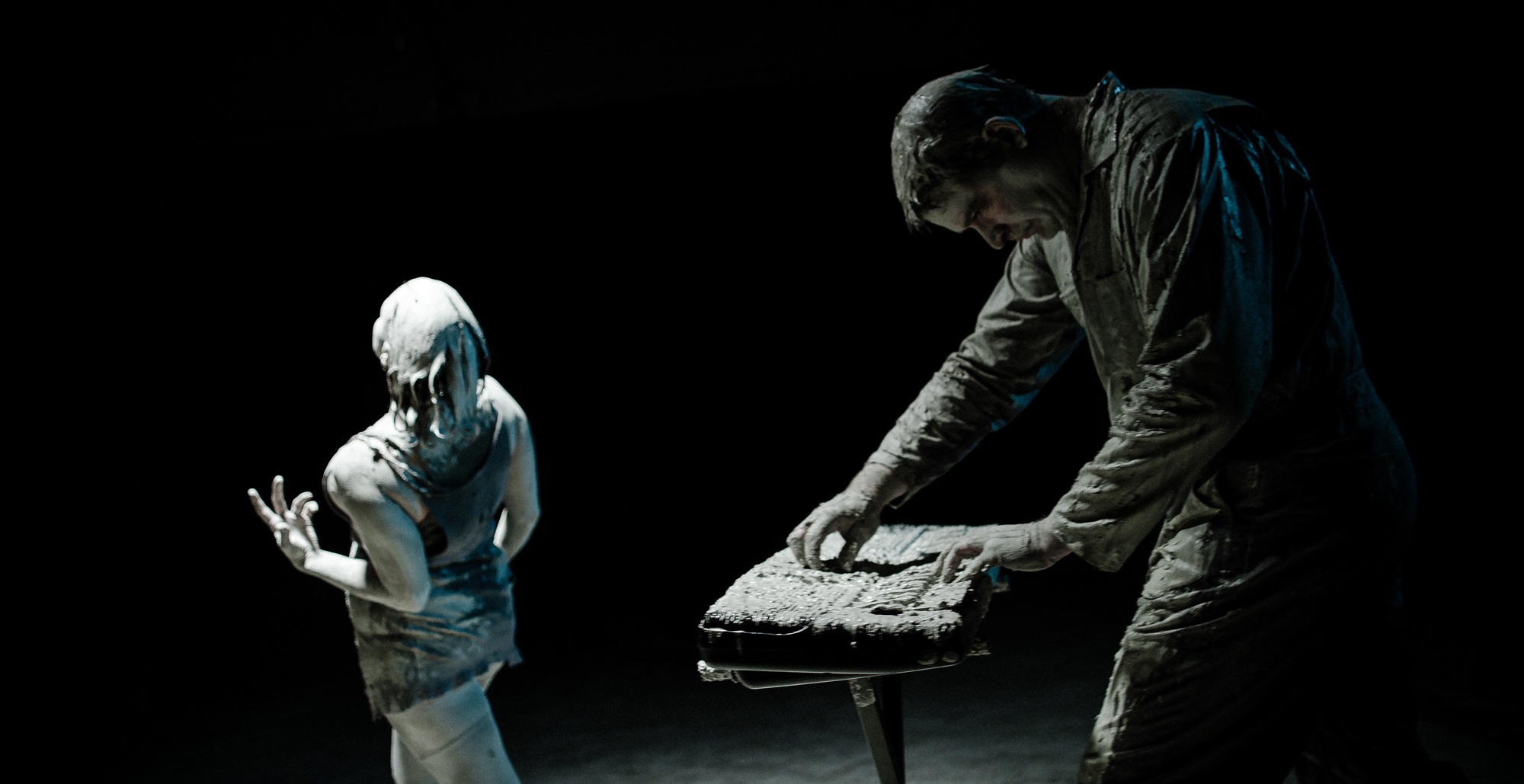 Man and woman covered in clay plays keyboards and dances in dark room lit from above.