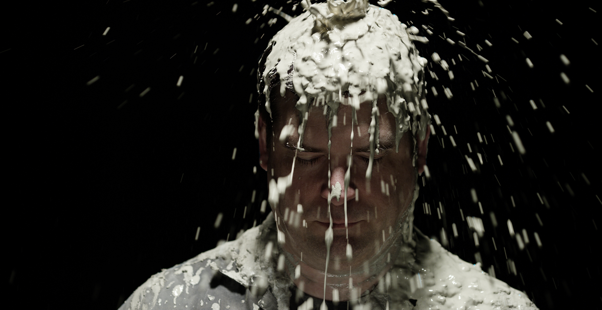 Man has mud pouring over face in spotlit dark room.