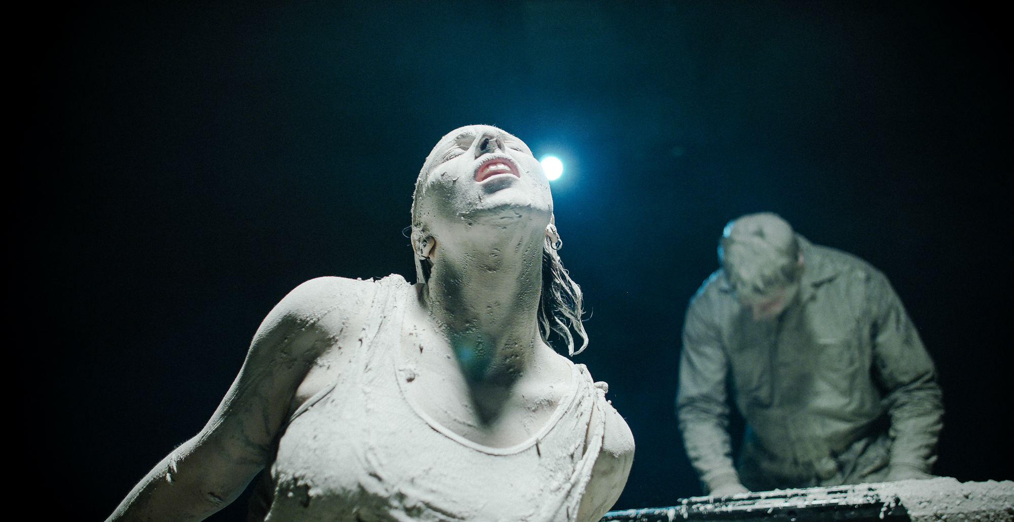 Man and woman covered in clay plays keyboards and dances in dark room lit from above.
